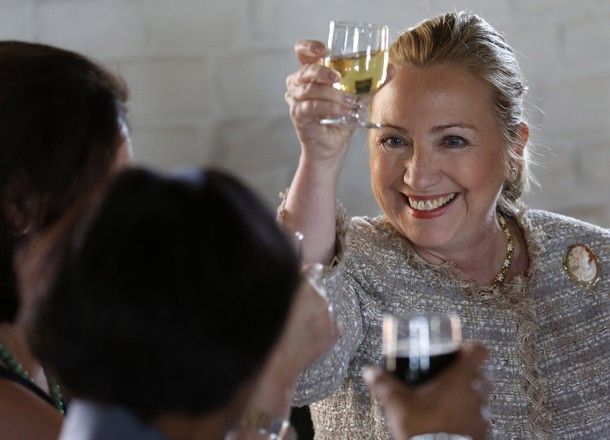 image of Hillary Clinton at a table for a meal, lifting her wine glass and smiling