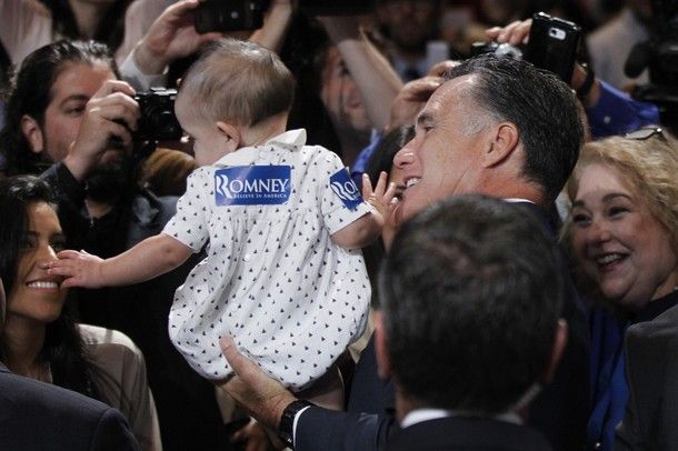 image of Mitt Romney holding a baby, who is plastered with Mitt Romney stickers, like a sack of potatoes over a crowd