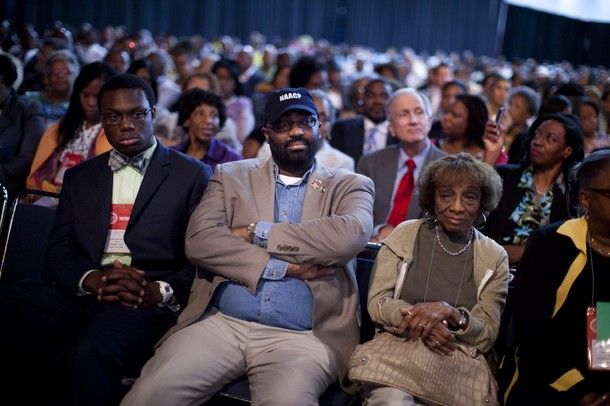 A man sits in the audience of Romney's NAACP address with his arms folded; to either side are a man and a woman, both looking unthrilled
