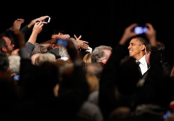 image of President Barack Obama in a crowd, visible between the raised arms of someone taking a picture of him on a mobile phone