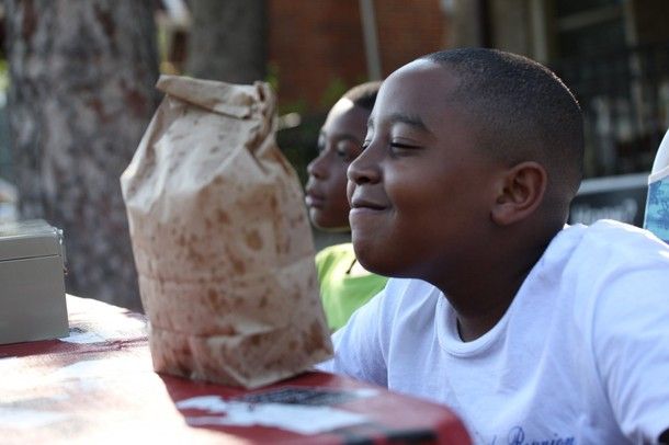 Joshua Smith, a young black boy, smiles while sitting with a bag of popcorn at his lemonade stand