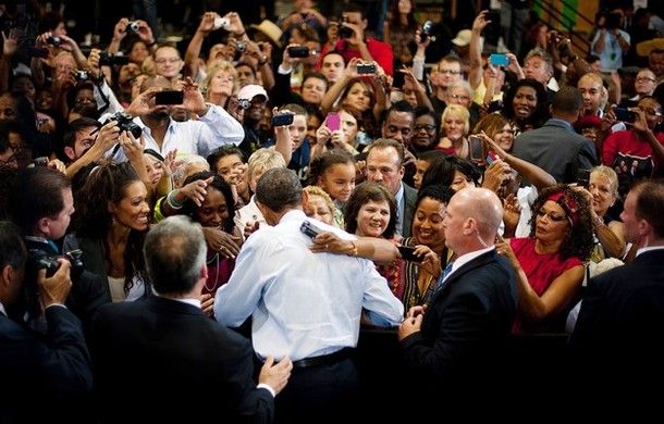 an image of a diverse crowd of people reaching for President Obama and taking pictures of him, as he hugs a woman at a campaign event, while Secret Security stands by