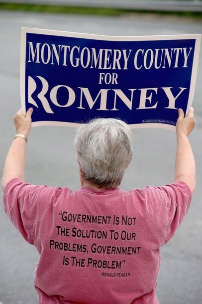 an image of a white woman from behind holding up a 'Montgomery County for Romney' sign; she is wearing a t-shirt emblazoned with annoyingly comma-spliced text reading: 'Government is not the solution to our problems, government is the problem. - Ronald Reagan'