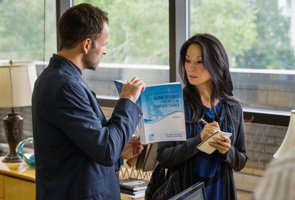 image of Sherlock Holmes (Jonny Lee Miller) and Joan Watson (Lucy Liu) from the latest episode of Elementary; they are both wearing grey blazers, and he is holding up a book titled 'Global Research in Theoretical Computer Science' from which she is taking notes