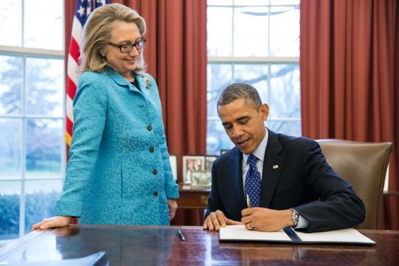 Obama signs paperwork while Clinton stands next to him