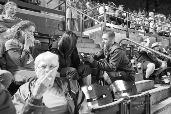 black and white image of a young white man proposing to a young white woman at a baseball game, while, in the foreground, an elderly white woman flips off the photographer who is obstructing her view