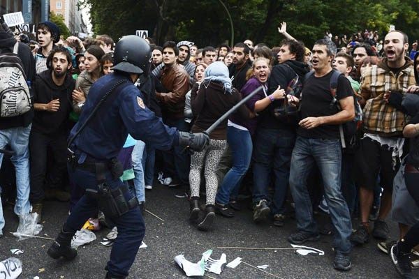 image of protestors in Spain cringing away from a police officer who is wielding a baton threateningly