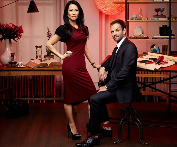 image of Lucy Liu and Jonny Lee Miller as Holmes and Watson; Liu is standing and wearing a deep red dress; Miller is seated and wearing a suit and deep red socks