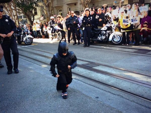 image of a little boy dressed as Batman running down a street in San Francisco, as people lining the sidewalks cheer him on