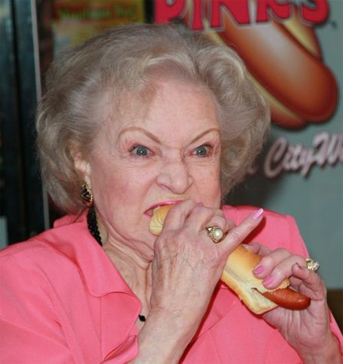 image of actress Betty White, an elderly white woman, biting ferociously into a hotdog with her pink-manicured pinky daintily raised