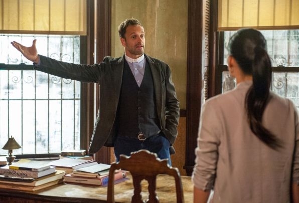 image of Sherlock and Joan standing beside a table; Sherlock is gesturing toward a window