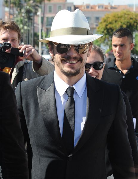 image of actor James Franco, a young white man, walking through a crowd at the Cannes Film Festival; he is smiling broadly and wearing a black suit, white fedora, and reflective sunglasses