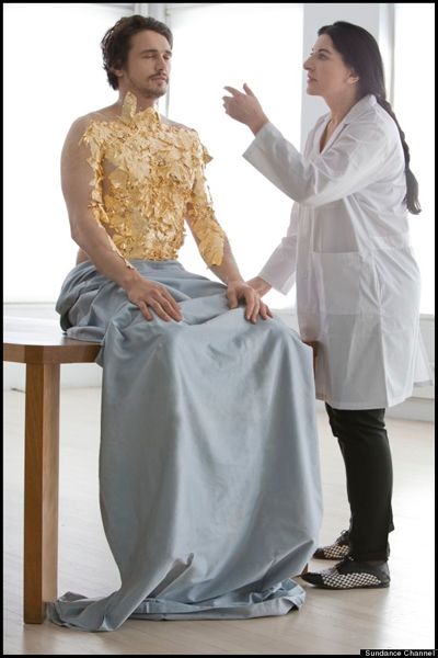 image of actor James Franco sitting naked on a table with a sheet draped over his lap and legs, his bare chest being covered in gold leaf by artist Marina Abramovic, who stands at his side in a white lab coat