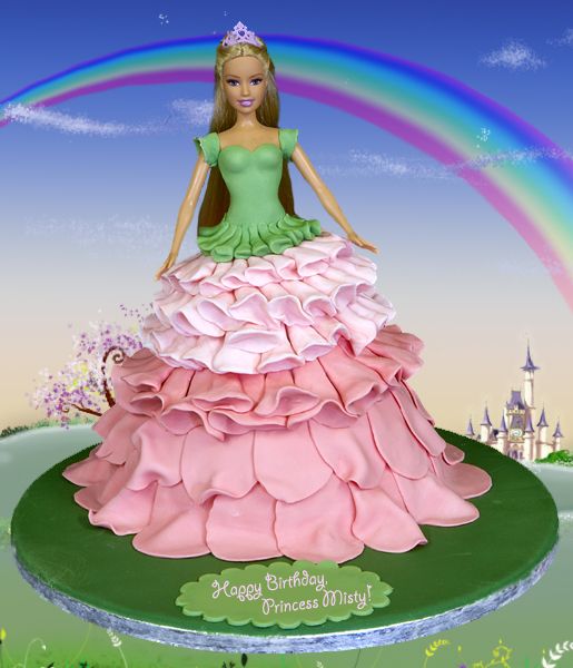 a Barbie cake in which the Barbie is wearing a pink and green princess gown and is standing in front of a fairyland with a sign labeled 'Happy Birthday, Princess Misty!'