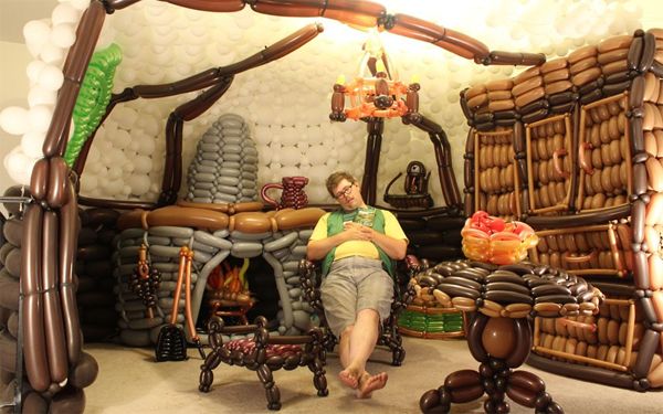 image of a mid-30s white man sitting in his living room which is covered in balloons arranged to make it look like a hobbit hole as described by J.R.R. Tolkien in his fantasy novels