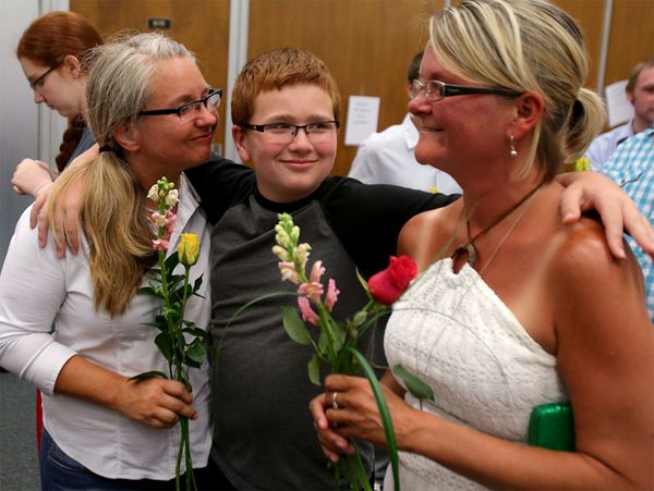 image of a young white boy with his arms around the shoulders of two older white women standing on either side of him, holding flowers as they wait to be married; he has a huge grin on his face