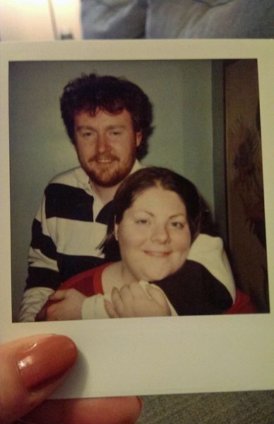 image of my hand holding an old photo of Iain and me; Iain is standing behind me with his arm around my shoulder and neck