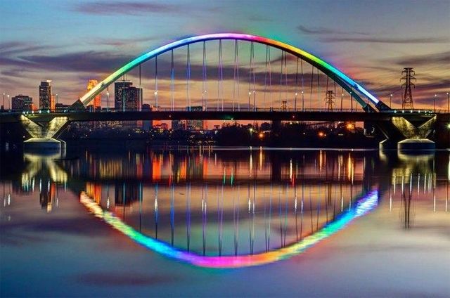 image of an arched bridge lit up with rainbow lights, and its reflection in the evening water