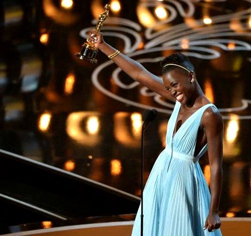 image of actress Lupita Nyong'o smiling and holding up her Oscar onstage after having won Best Supporting Actress