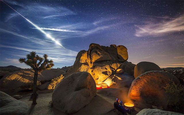 image of two people sitting at a fire at a desert campsite, as a meteor streaks across the night sky