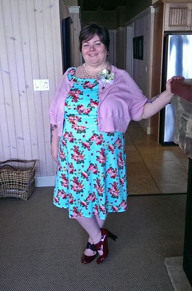 image of me standing in a cottage, wearing a turquoise dress with a rose print, a pink short-sleeved cardigan, pink pearls, and red patent shoes
