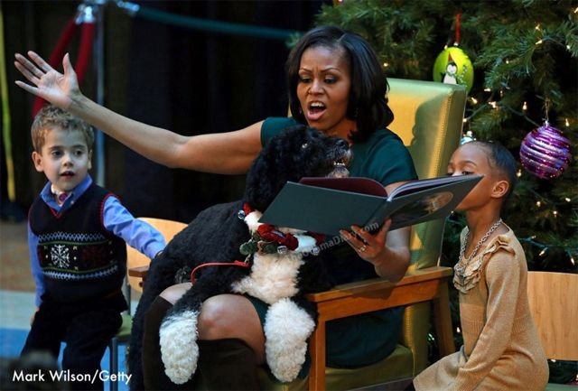 First Lady Michelle Obama gestures with a sweeping arm and expressive face while reading from a children's book, with a little boy and little girl at her sides, and her dog on her lap