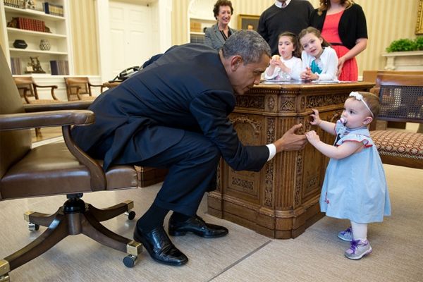image of President Obama sitting at his desk in the Oval Office, reaching over to let a little baby girl, who appears to be white, hold his finger as she balances to stand, while her older sisters look on