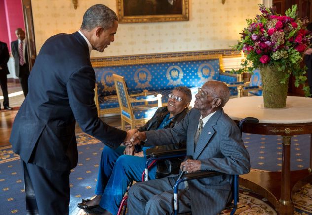 image of President Barack Obama leaning over to shake the hand of an elderly black man seated in a wheelchair; beside him sits an elderly black woman, also seated in a wheelchair
