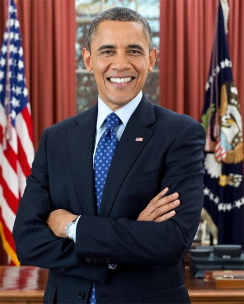 image of President Barack Obama in the Oval Office, standing and smiling with his arms crossed; he is wearing a blue suit jacket, white shirt, and blue tie with lighter blue dots