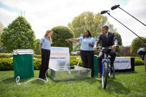 image of President Obama intently pedaling a bicycle connected to a water filtration system as its two creators, a black teenage girl and a white teenage girl, explain the science