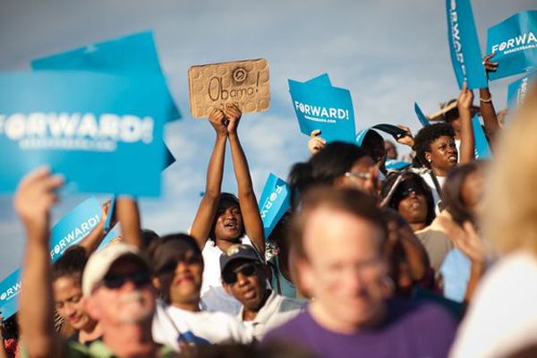 image of a young black woman at a campaign rally holding up a handmade cardboard sign with 'Obama!' written on it alongside a spray-painted Obama campaign logo