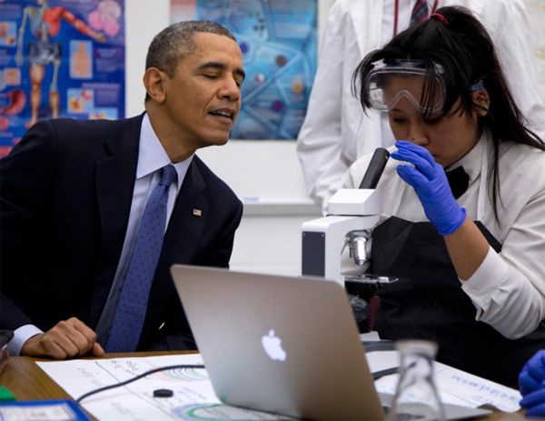 President Obama leans over the examine what a young woman of color is viewing with a microscope