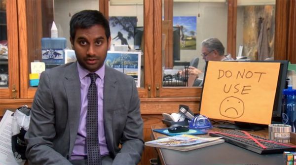 image of Tom (Aziz Ansari) sitting at his desk next to his computer monitor, which has been covered by a sign reading: 'Do Not Use' followed y a frowny-face