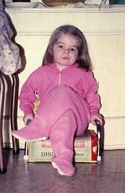 image of me as a tiny little kid, clad in pink footie pajamas and sitting with my legs crossed in a child's chair, making a contemptuous expression