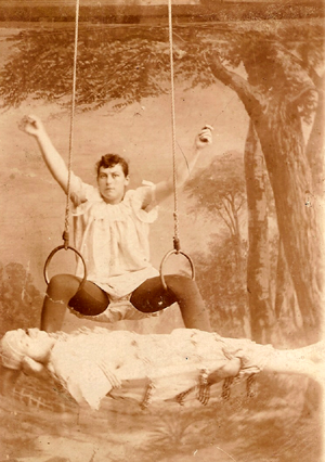 image of a fat white woman balancing in a sitting position with her legs between two acrobatic hoops and her arms in the air; across the tops of her feet is balanced a person who looks to be clad as a mummy