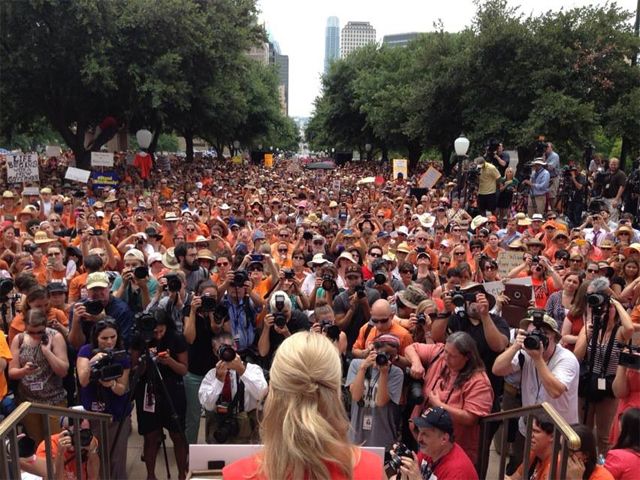 image of Wendy Davis speaking at the rally in Texas today; we see the back of Davis' head, with a gaggle of photographers in front of her, and a crowd stretching into the distance