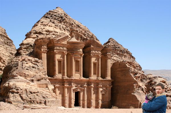 actor Tom hardy, holding a grey pit bull puppy who is licking its nose, stands in front of the monastery at Petra, a building carved into the side of a mountain