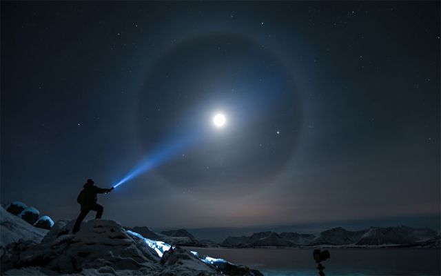 image of a man on a hilltop holding out a flashlight so it looks as though he's creating a halo around the moon