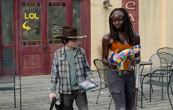 image of Carl and Michonne walking away from a cafe; Carl is holding a picture and Michonne is holding a multi-colored cat figurine; in the background, a zombie's face is seen pressed against the window; I have added text reading 'LOL' with an arrow pointing to the zombie's face