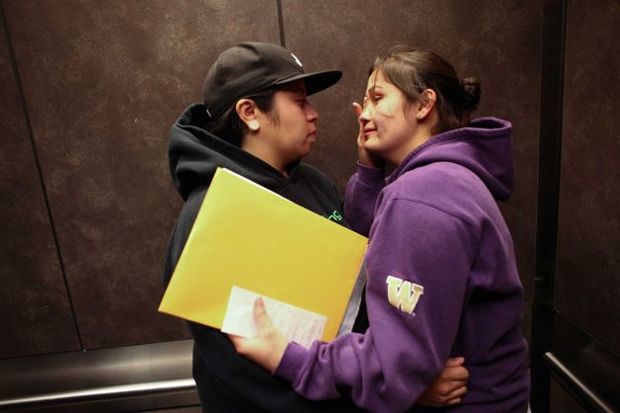 image of two young women, one a woman of color and one who appears to be white, hugging each other in an elevator while holding a newly acquired marriage license, and wiping away tears