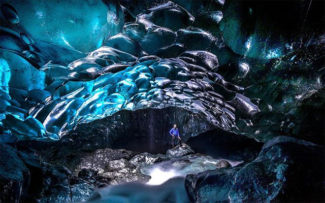 image of a thin white man standing in the middle of a beautifully blue-glowing glacial cave