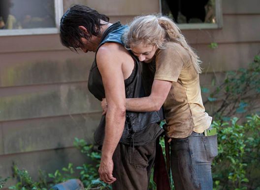 image of Daryl and Blonde Girl standing outside a cabin; his head is hanging forward and she's hugging him from behind