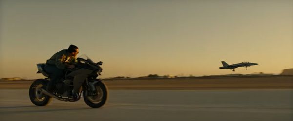 Maverick races his motorcycle against an F/A-18 Hornet as it takes off from a runway in TOP GUN: MAVERICK.